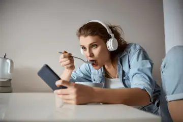 Młoda kobieta w słuchawkach je płatki, patrząc w telefon. Czy to objaw fonoholizmu?