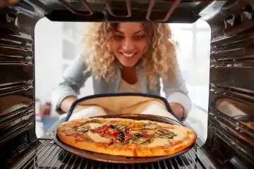Uśmiechnięta kobieta wyjmuje z piekarnika pyszną, gorącą pizzę. Właśnie skończyła piec pizzę i jest gotowa do jedzenia.