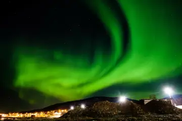 Niesamowita zorza polarna w Polsce rozświetla nocne niebo. Zielone pasma tańczą nad oświetloną wioską i wzgórzami.