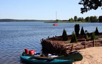 Dwie kajaki na piaszczystej plaży w Swornegaciach na Kaszubach. W oddali czerwona łódka i jezioro.