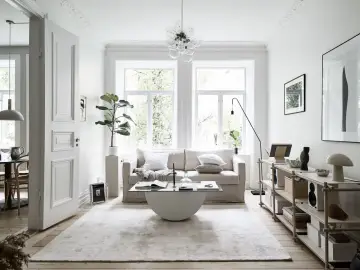 Salón con sofá beige, mesa de centro de cristal y alfombra clara. Decoración minimalista y plantas, creando un ambiente de feng shui.