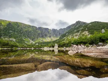 Spokojne, górskie jezioro odbija skaliste zbocza i chmury. W oddali widać skalne formacje, a brzegi porasta bujna zieleń. To idealne miejsce na podziwianie piękna przyrody, jakbyśmy patrzyli na śnieżne stawki.