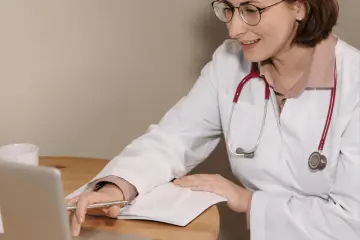 Lekarka w białym kitlu z czerwonym stetoskopem analizuje dane na laptopie i notuje w zeszycie. Dowiaduje się, jak zostać lekarzem medycyny pracy.