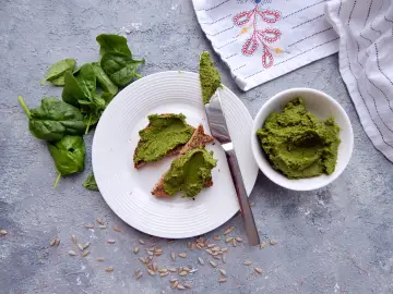 Pesto ze szpinaku na grzankach, obok miseczka z pastą i świeże liście szpinaku.