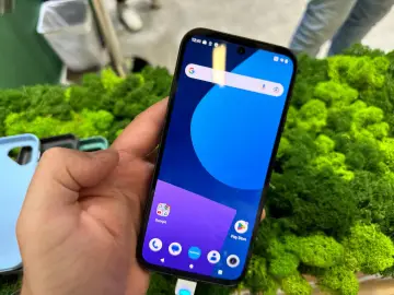 Dłoń trzyma smartfon z kolorową tapetą. W tle zielony mech i kilka etui do różnych **marek telefonów**.