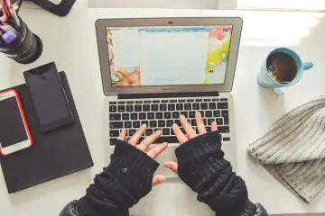 Ręce na klawiaturze laptopa, obok telefon, kubek herbaty i czapka. Jak wyłączyć tryb recenzji w Wordzie?