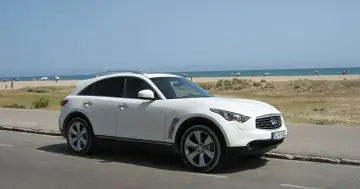 Białe infiniti auto zaparkowane przy plaży, z widokiem na morze i ludzi odpoczywających na piasku.