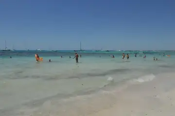 Słoneczny dzień na plaży Es Trenc. Ludzie kąpią się w turkusowej wodzie, a na horyzoncie widać żaglówki.