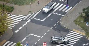 Na skrzyżowaniu równorzędnym dwa samochody zbliżają się do pasów. Jeden biały, drugi srebrny.