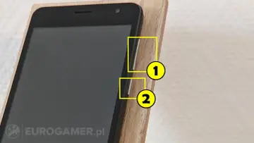 Zbliżenie na czarny ekran telefonu w drewnianej podstawce. Dwa żółte kółka z cyframi 1 i 2 wskazują przyciski boczne.