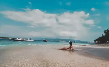 Kobieta w bikini siedzi w płytkiej wodzie na plaży, patrząc na morze z łodziami. To prawdziwy raj na ziemi.