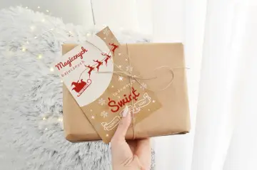 Ręka trzyma paczkę z papieru pakowego, ozdobioną kartką z Mikołajem i reniferami. To z pewnością fajne prezenty na święta!