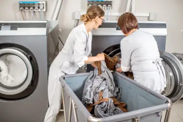 Pracownice pralni chemicznej w białych fartuchach wkładają pranie do profesjonalnych pralek. To pokazuje, dlaczego profesjonalne pralnie chemiczne nie używają zwykłych proszków do prania – potrzebują specjalistycznych środków.