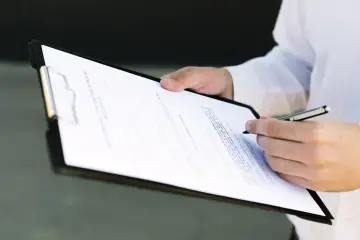 Dłoń podpisuje dokument, być może umowę kupna-sprzedaży nieruchomości. W tle widać fragment białej koszuli. Warto sprawdzić, czy nie ma tam ukrytych informacji o vat-r.