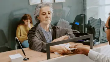 Starsza pani odbiera pieniądze w banku, być może związane z nowym wiekiem emerytalnym w Polsce.