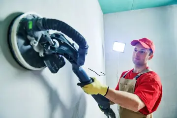 Pracownik w czerwonej koszulce i ogrodniczkach używa szlifierki do ścian, by uzyskać idealną strukturę z gładzi.