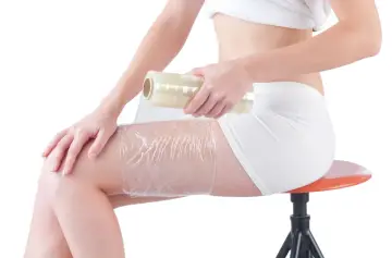 Kobieta owija udo folią spożywczą, przygotowując się do zabiegu body wrapping. Widoczne efekty zabiegu to m.in. wygładzenie skóry.