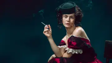 Kobieta w stylu Anny Kareniny, w eleganckiej sukni i kapeluszu z woalką, pali papierosa. Dym unosi się wokół jej twarzy.