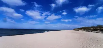 Pusta, piaszczysta plaża nad polskim morzem. Błękitne niebo z białymi chmurami, ciemne morze i wydmy porośnięte trawą.