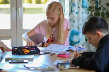 Dzieci odrabiają lekcje. Dziewczynka z blond włosami przegląda zeszyt, chłopiec pisze. Wygląda na to, że nie ma zadania domowego.