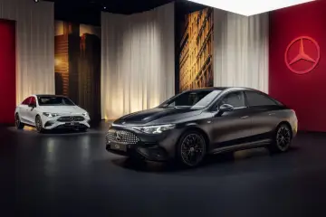 Dwa modele Mercedes CLA 2025: jeden biały, drugi grafitowy, prezentowane na tle miejskiej panoramy i czerwonej ściany z logo marki.