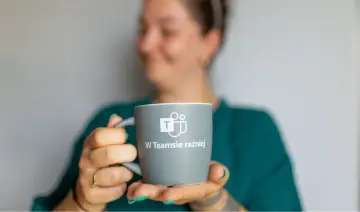 Kobieta trzyma kubek z logo Microsoft Teams i napisem "W Teamsie raźniej". Idealny do kawy podczas pracy zdalnej lub rozmów z helpdeskiem.