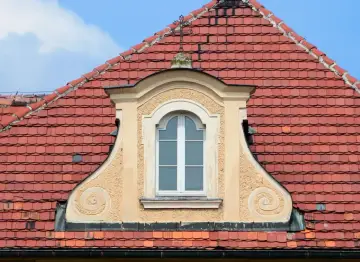 Eleganckie, małe okienko w dachu z ozdobnymi wolutami, otoczone czerwonymi dachówkami.