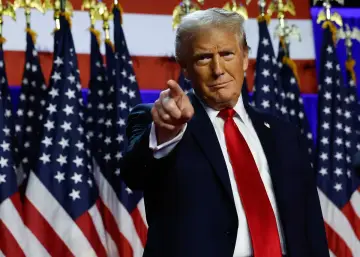 Donald Trump, prezydent USA, wskazuje palcem na widza, stojąc przed amerykańskimi flagami.