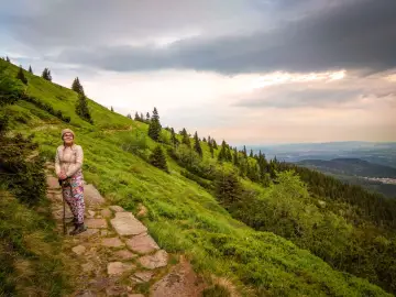 Kobieta na szlaku w górach ze śnieżką. Zielone zbocza, kamienna ścieżka i chmury na niebie.