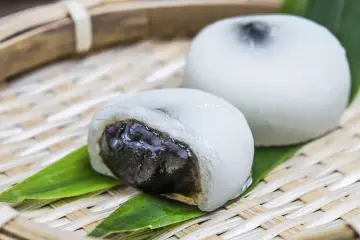 Dwa białe mochi, jedno przekrojone, ukazujące ciemne nadzienie. Podane na bambusowej tacce z liśćmi.
