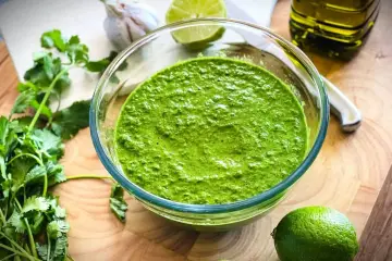 Frisch zubereitetes mojo verde in einer Glasschüssel, umgeben von Koriander, Knoblauch, Limetten und Olivenöl.