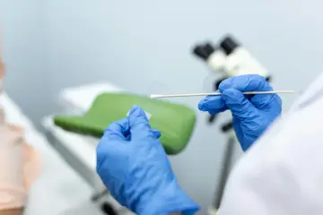 Ręce w niebieskich rękawiczkach przygotowują próbkę do badań cytologia wyniki. W tle mikroskop i fotel ginekologiczny.