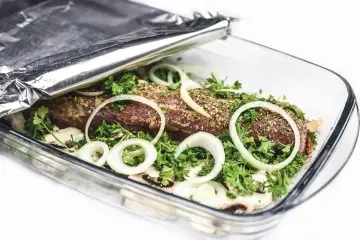 Mięso w naczyniu żaroodpornym, przykryte folią aluminiową, z cebulą i ziołami.