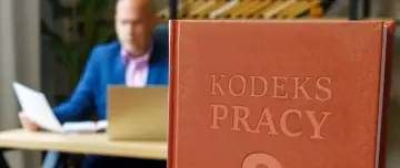 Mężczyzna w garniturze przegląda dokumenty przy biurku, obok stoi oprawiony "Kodeks Pracy", który przypomina o jego uchwaleniu.