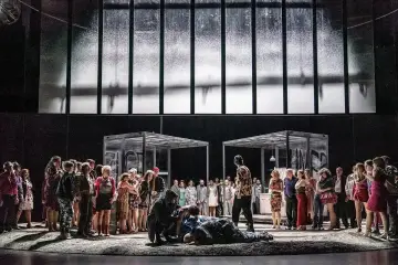 Scena z opery Paderewskiego: tłum ludzi na scenie, dwie szklane konstrukcje, na pierwszym planie leżący mężczyzna.