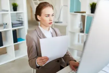 Młoda kobieta w biurze, przygotowująca się do szkolenia wstępnego BHP, analizuje dokumenty przy komputerze.