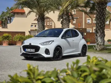Biała Mazda 2 Hybrid zaparkowana przed budynkiem z palmami. Stylowy miejski samochód gotowy na podróż.