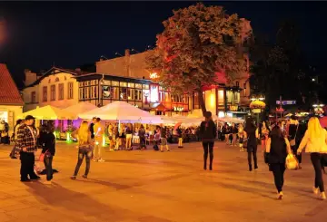 Tętniący życiem wieczór w Sopocie. Ludzie spacerują po oświetlonym placu, gdzie znajdują się restauracje i kawiarnie pod białymi parasolami.