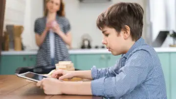 Chłopiec gra na tablecie, podczas gdy mama w tle zastanawia się, jak usunąć telefon z Family Link.