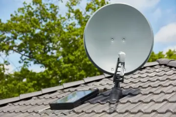 Srebrna antena satelitarna na szarym dachu, obok okna dachowego. W tle zielone drzewa i błękitne niebo.