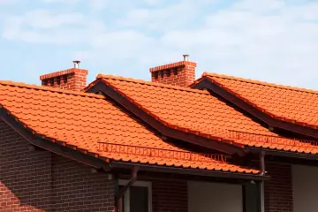 Pomarańczowy dach z dachówki ceramicznej, z ceglanymi kominami i brązowymi rynnami, pod błękitnym niebem.