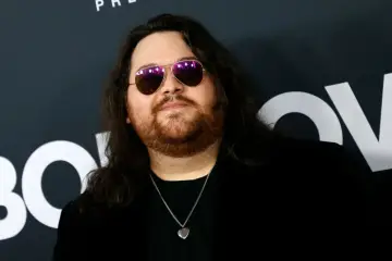 Wolfgang Van Halen w okularach przeciwsłonecznych i z naszyjnikiem w kształcie serca, na tle logo HBO.
