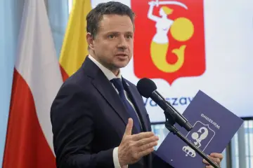 Prezydent Warszawy w garniturze przemawia przy mikrofonie, trzymając teczkę z herbem miasta.