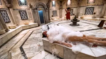 Kobieta w pianie podczas rytuału hammam. W tle inna kobieta w czerwonym ręczniku.