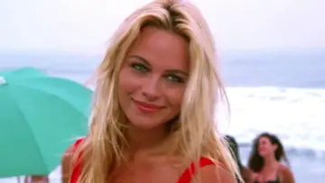 Słoneczny patrol obsada: blond ratowniczka w czerwonym stroju kąpielowym uśmiecha się na plaży, w tle ocean i parasol.
