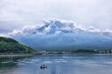 Majestatyczny Fudżi, otoczony chmurami, góruje nad jeziorem. W oddali most, a na wodzie łódka z wędkarzami. Zastanawiasz się, ile się leci do Japonii?