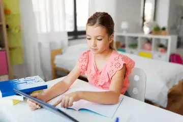 Dziewczynka korzysta z tabletu podczas nauczania domowego w podstawówce. Na biurku leżą książki i zeszyt.