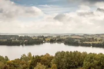 Spokojne **chańcza jezioro** otoczone zielonymi lasami i pagórkami. Na horyzoncie widać domy i pola.