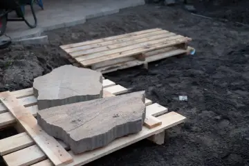 Płaskie kamienie na ścieżkę leżą na drewnianych paletach, gotowe do ułożenia.