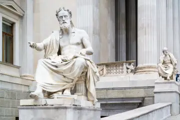 Kamienna rzeźba siedzącego mędrca, przypominającego Heroda, z wieńcem laurowym na głowie, na tle monumentalnej budowli.